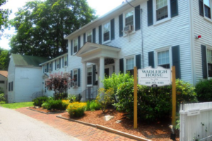 A white colonial style house with green shutters; the picture is taken from a sharp angle and shows the front of the house, the portico, and a brick walkway with landscaping.