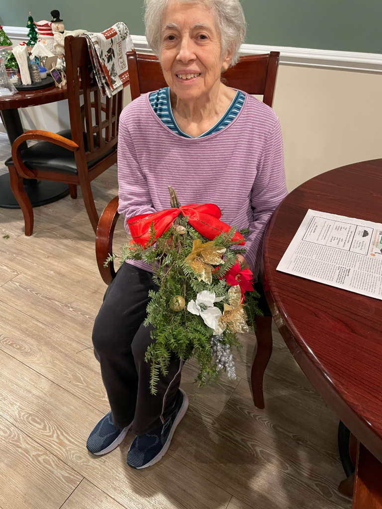 A resident is seated, holding a wreath they made, smiling.